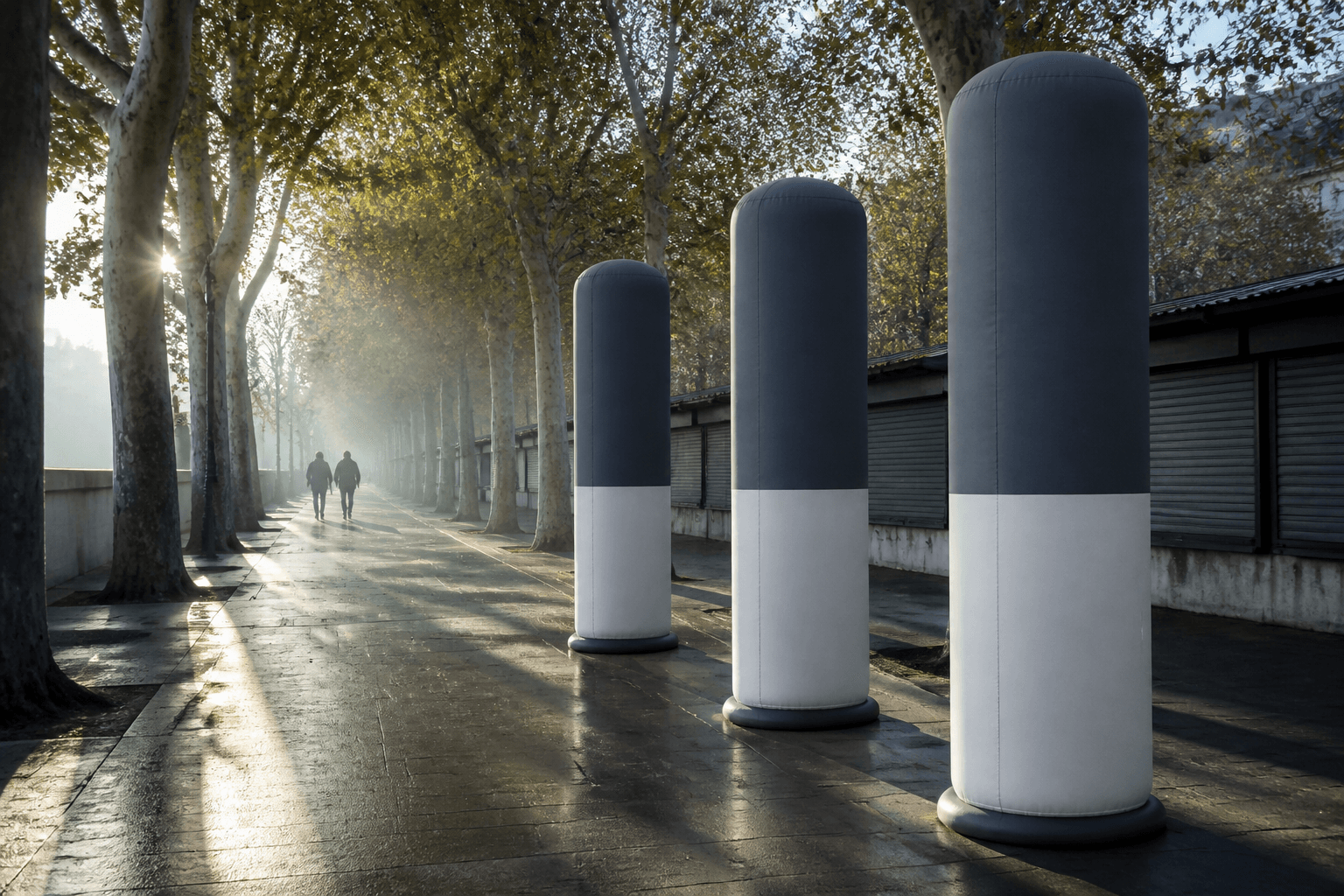 Three slate grey and off-white inflatable advertising columns along a tree-lined promenade